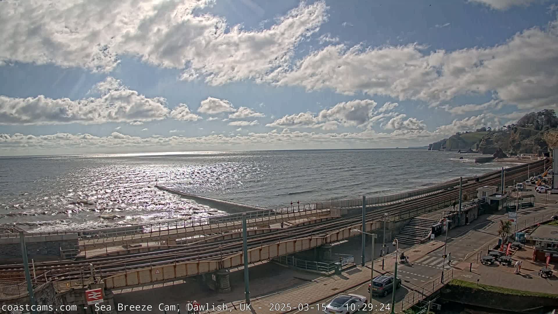 A partly cloudy day shows a coastal scene with a train track running alongside a beach and ocean with small waves.