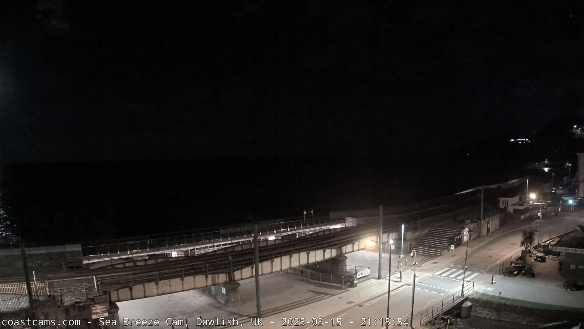 A nighttime view of a coastal town, showing a train trestle over a road leading to a dark ocean under a clear sky.