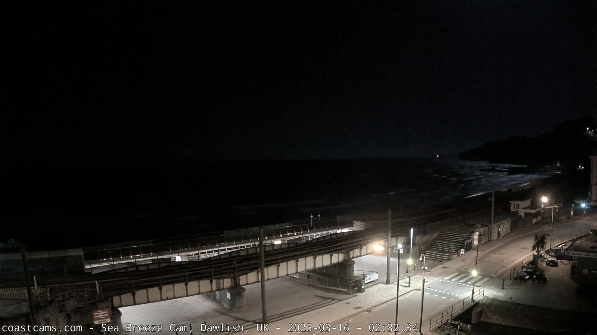 A nighttime coastal scene shows a dark ocean with waves crashing on the shore, a street with a pedestrian crossing, and a railway bridge extending over a walkway.