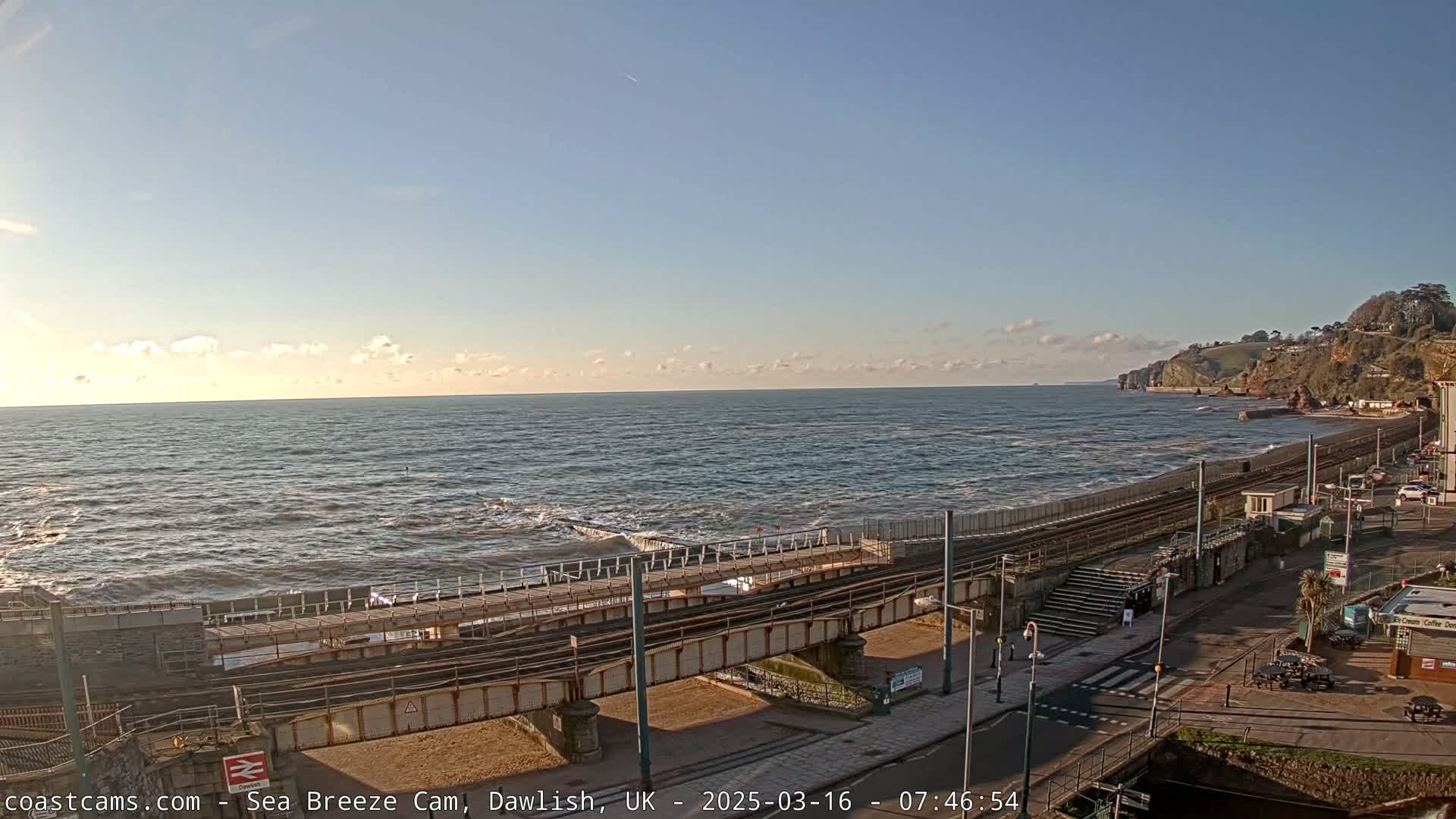 A mostly clear, sunny day shows a choppy sea meeting a coastal town with train tracks running alongside the shore.