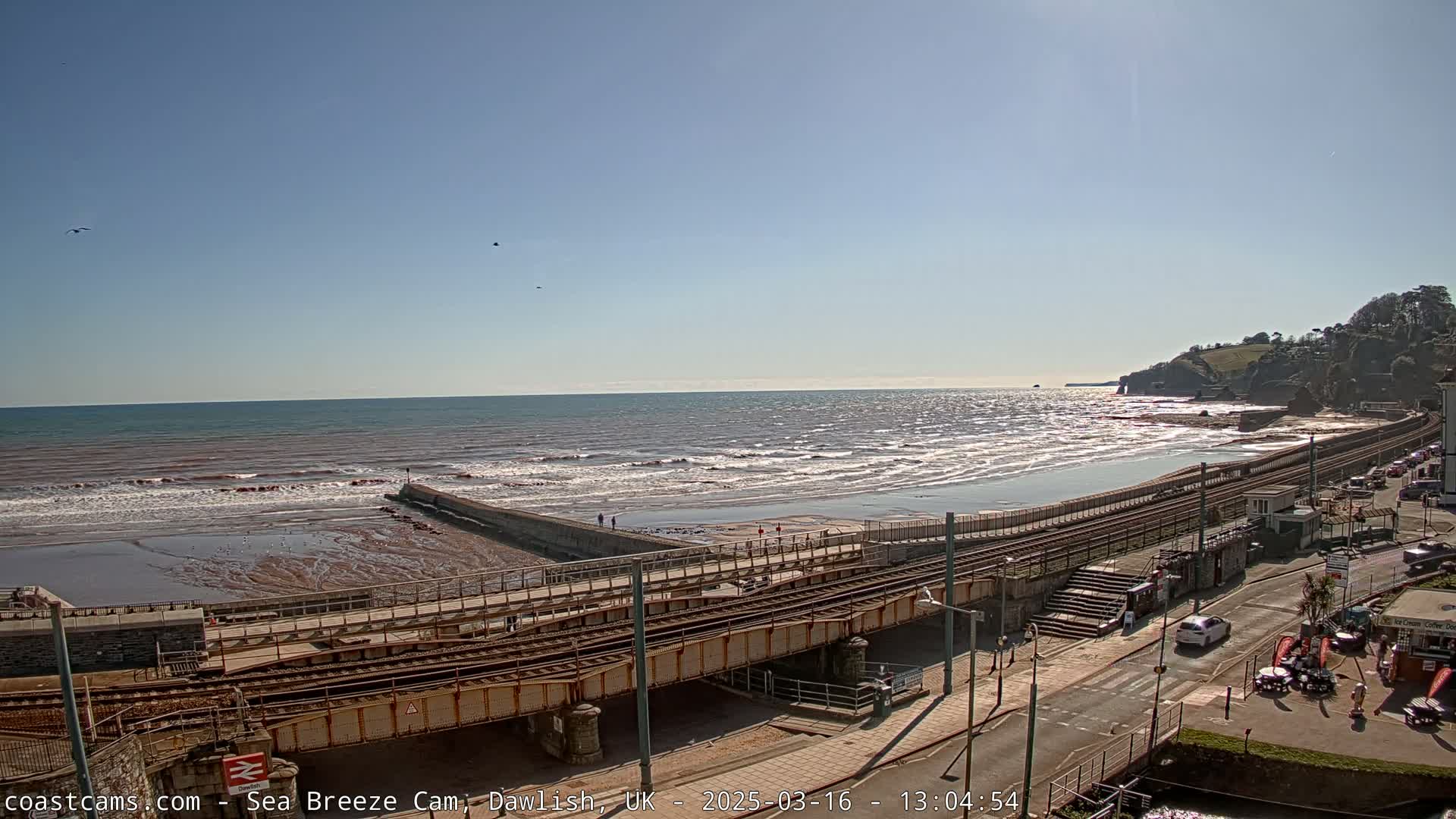 A sunny day at a beach with a railway line running along the shore, showing moderate waves and a few people walking near the water.