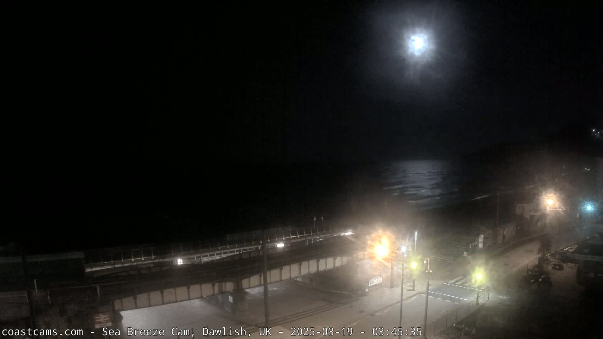 A nighttime view of a moonlit ocean with waves gently lapping the shore, seen from above a road and railway bridge.