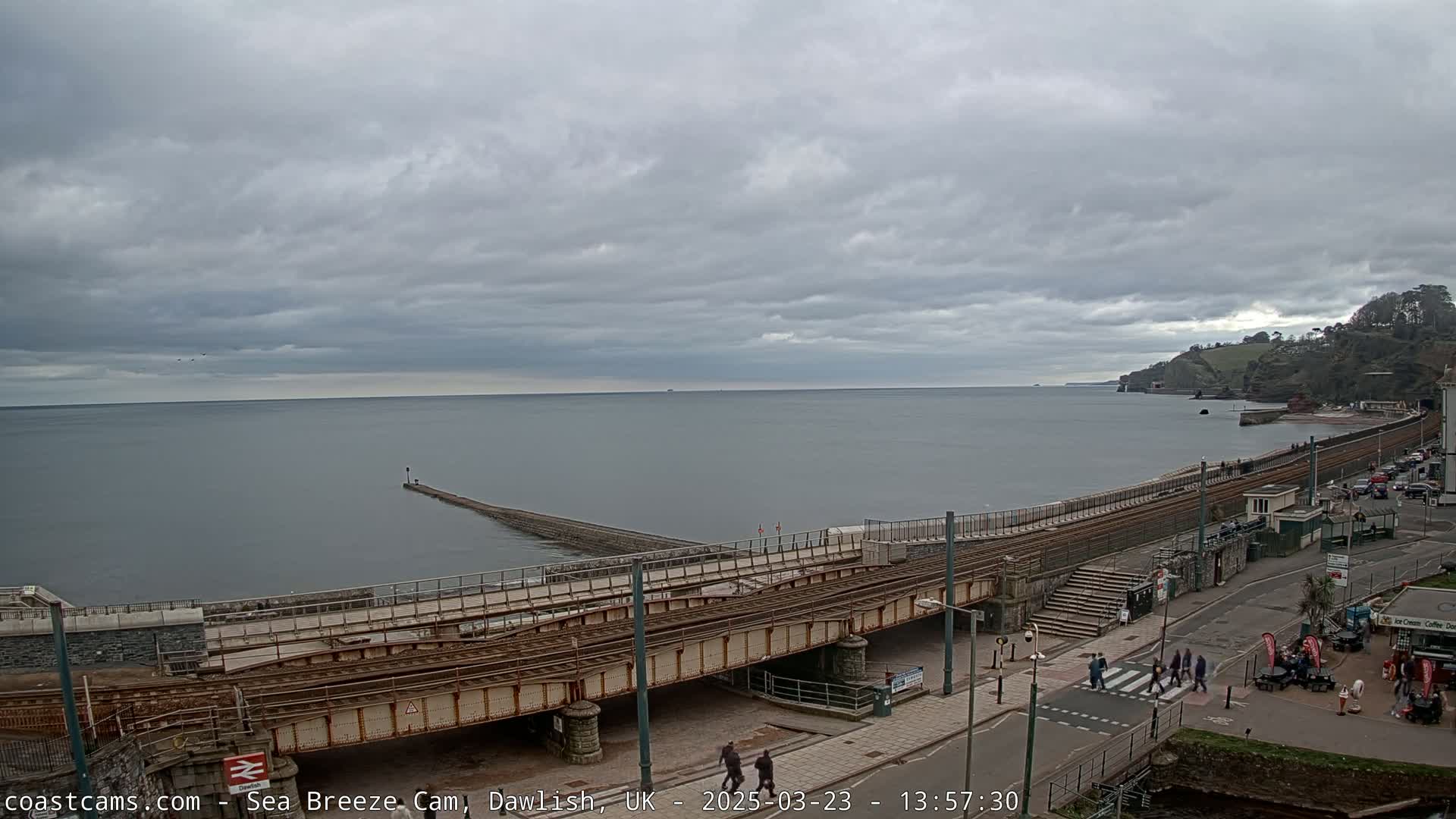 A mostly cloudy day overlooks a calm sea with a breakwater and railway tracks leading to a coastal town.