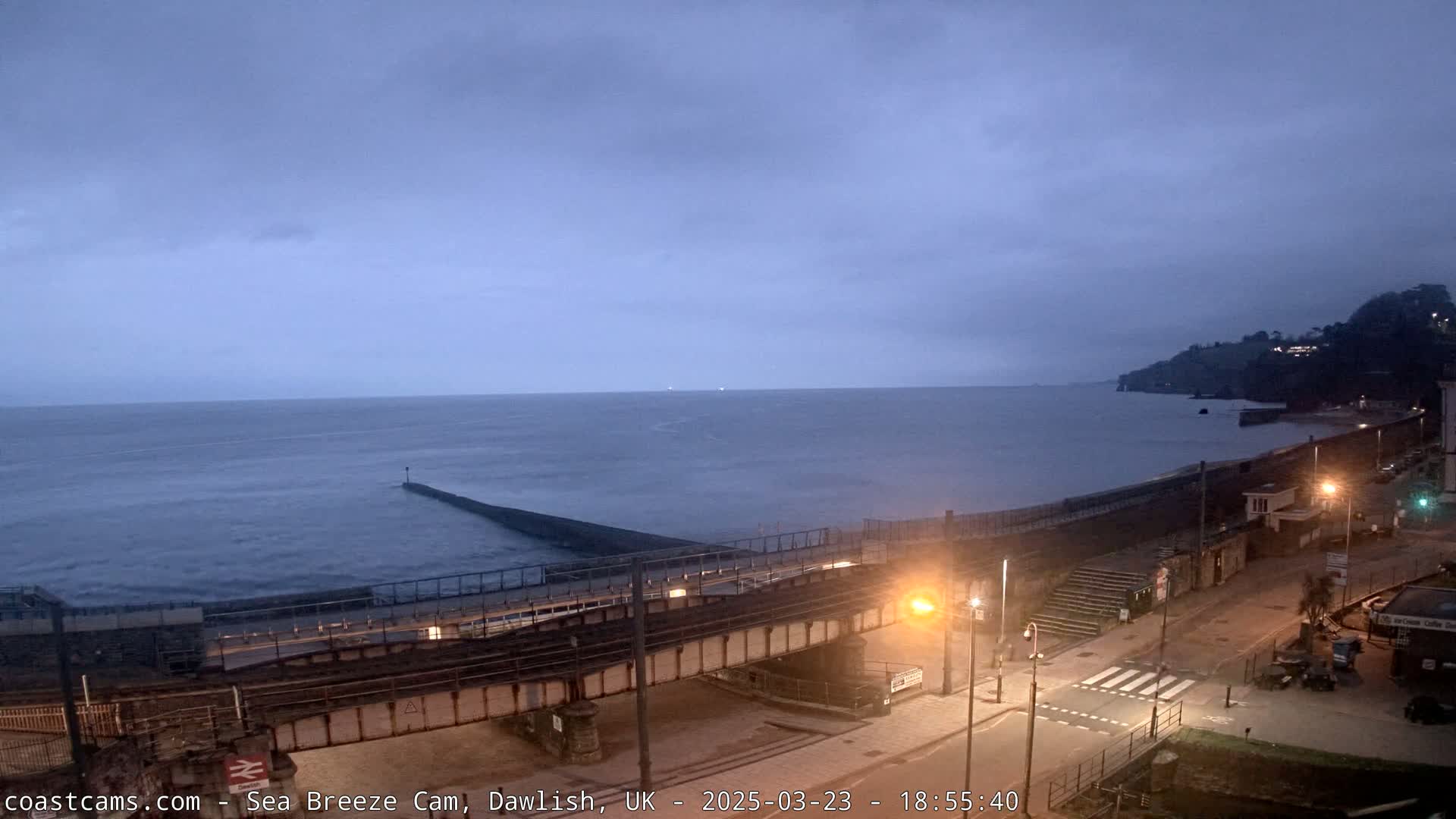 A dark, overcast coastal scene shows a sea wall, a railway bridge, and a road with a pedestrian crossing under a dusky sky.