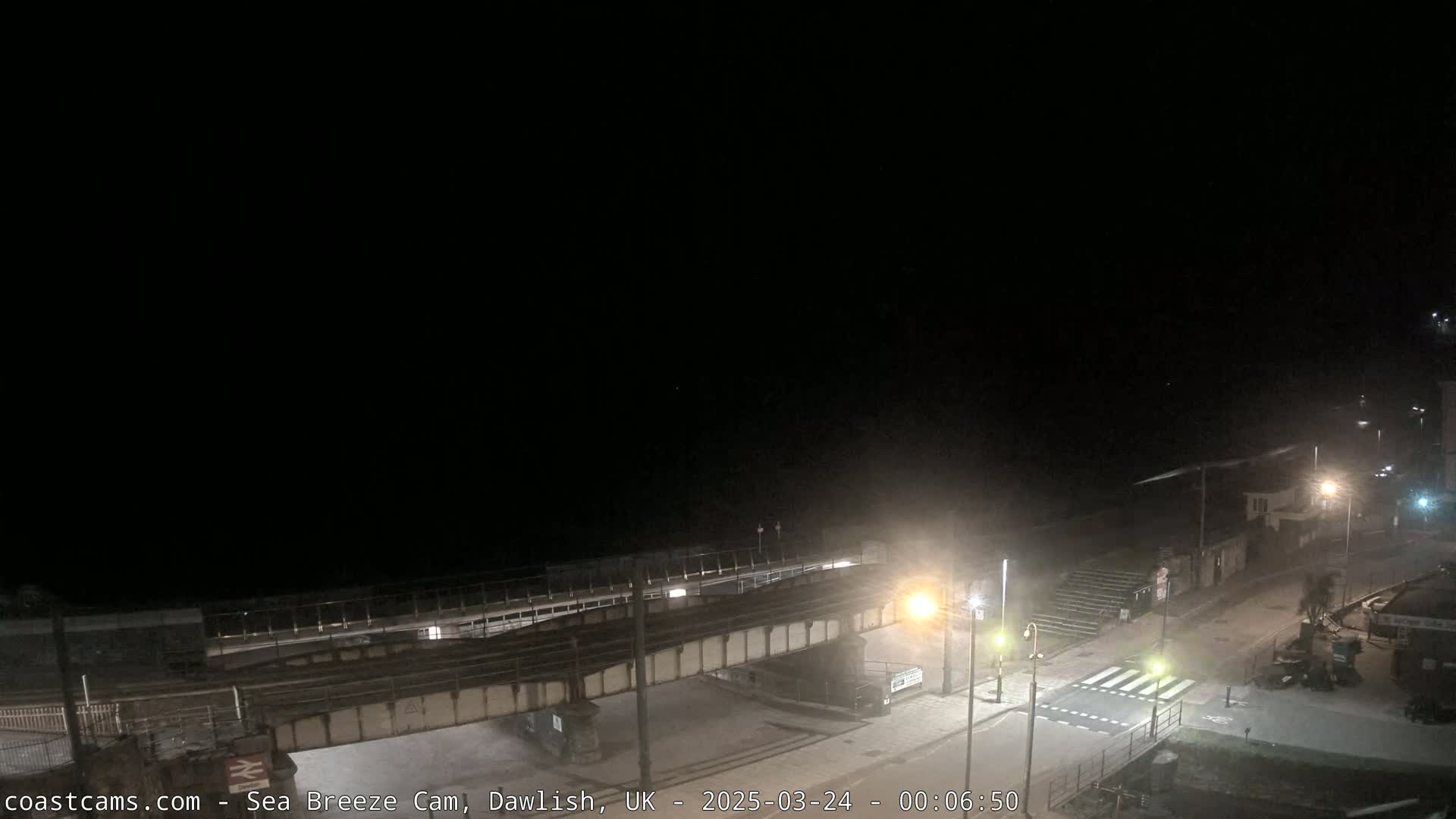 A nighttime view of a coastal town, showing a railway bridge over a road leading to a beach under a dark sky.