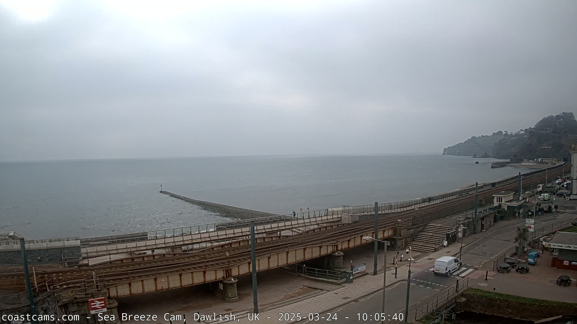 A coastal railway line crosses a bridge over a road next to a calm, gray sea under an overcast sky.