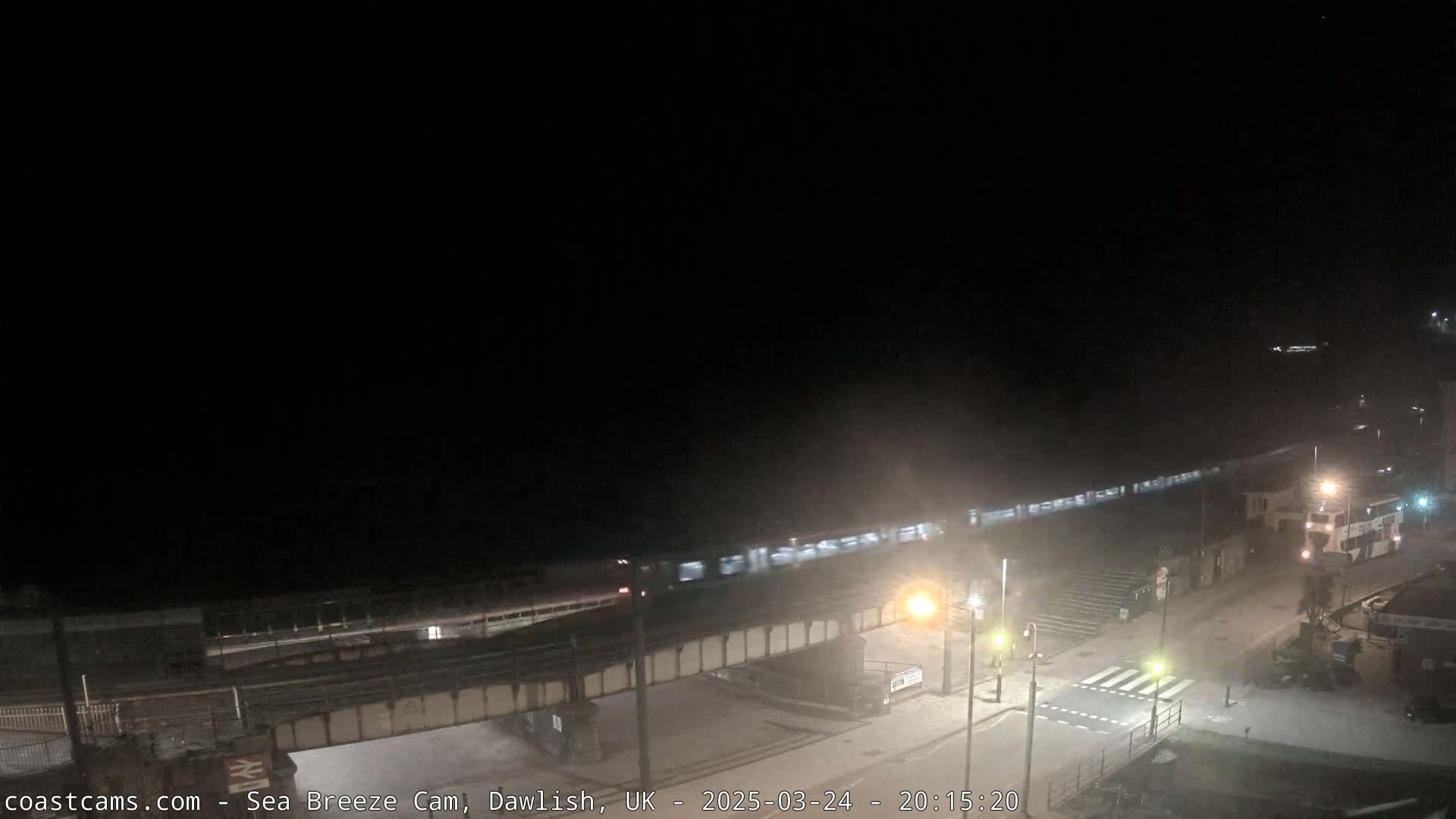 A train passes over a bridge at night near a coastline under dark, cloudy skies.