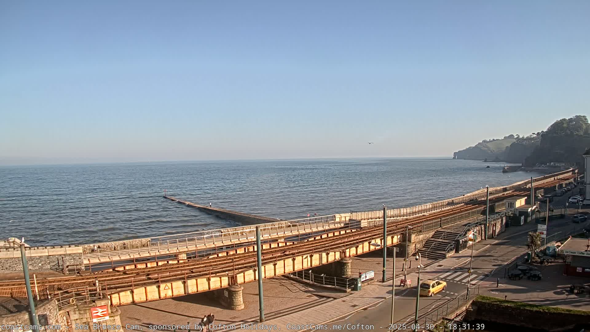 A coastal scene on a sunny day shows a railway track running alongside a sea wall, with a calm sea extending to a distant cliff.