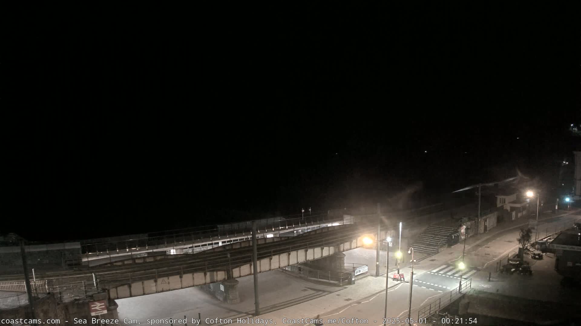 A nighttime view of a seaside town, showing a railway bridge over a road leading to a beach under a dark, clear sky.