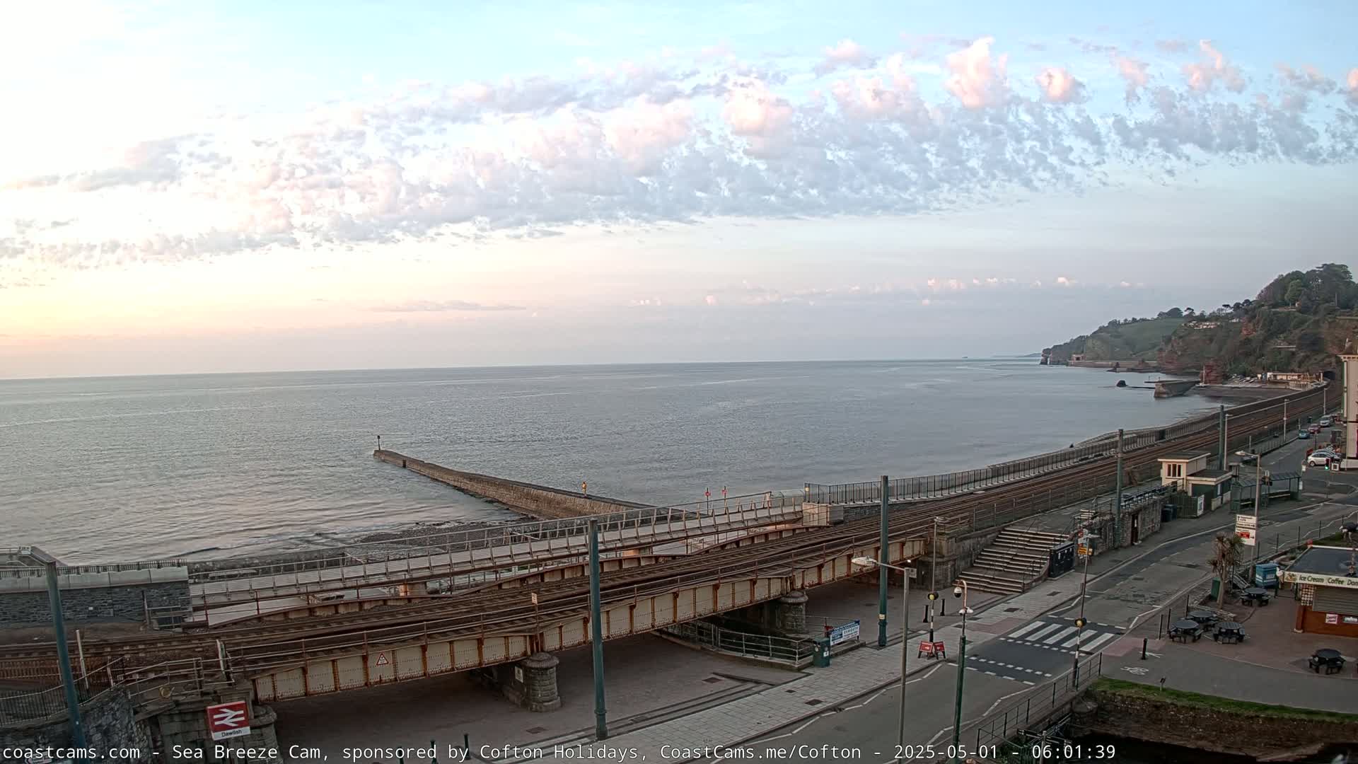 A partly cloudy sunrise illuminates a coastal scene featuring a railway crossing over a road leading to a sea wall and shoreline.