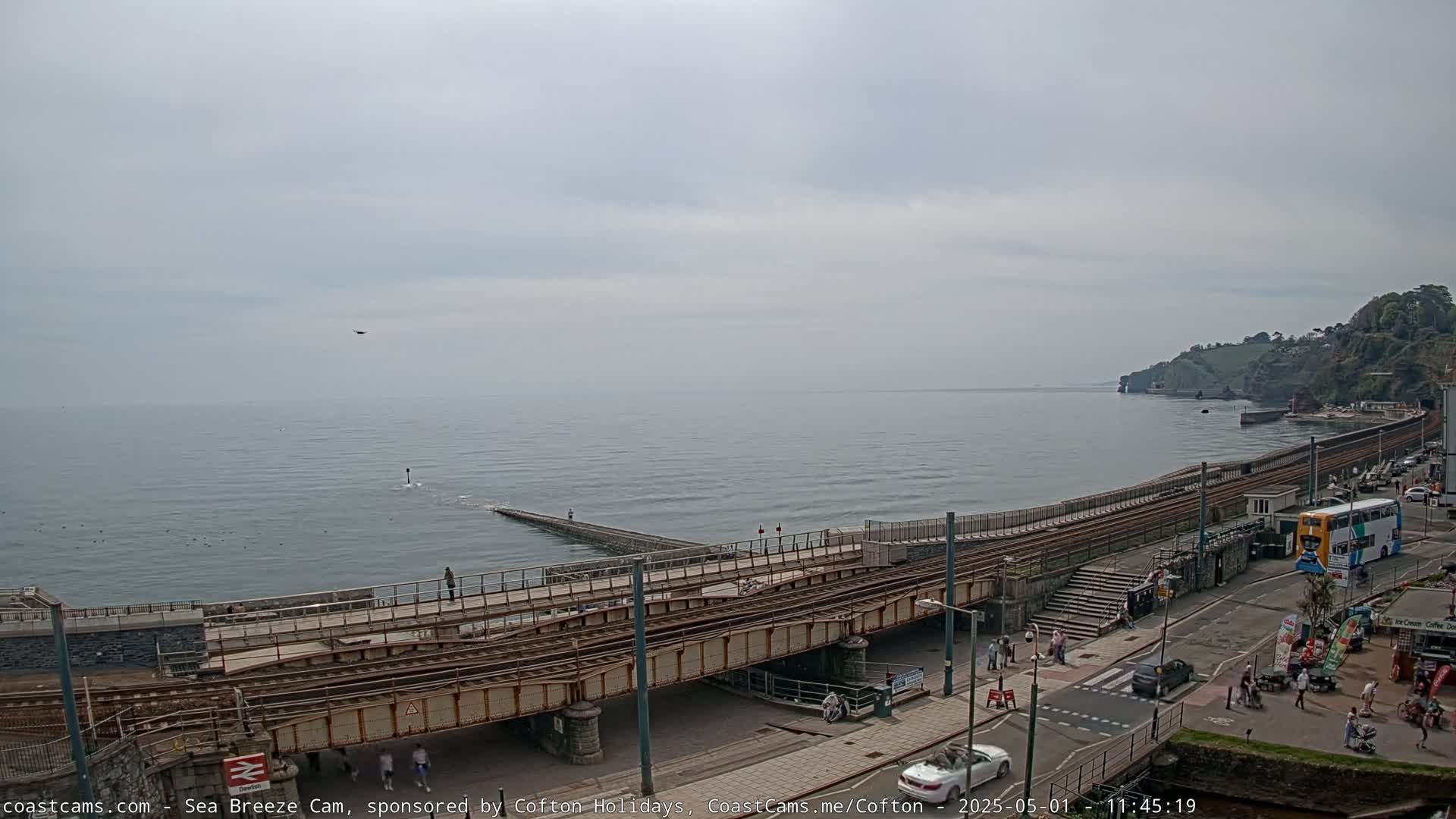 A coastal railway line crosses a road above a seawall under an overcast sky, with a bus and pedestrians visible on the road.