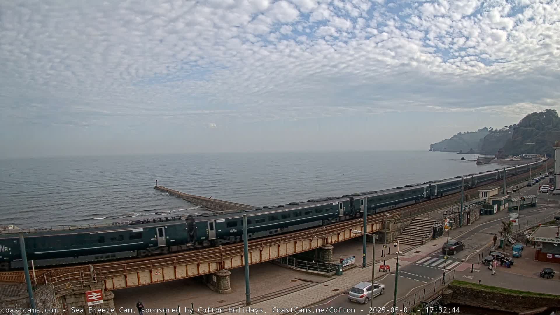 A long, dark-green passenger train travels along a coastal railway line under a mostly cloudy sky.