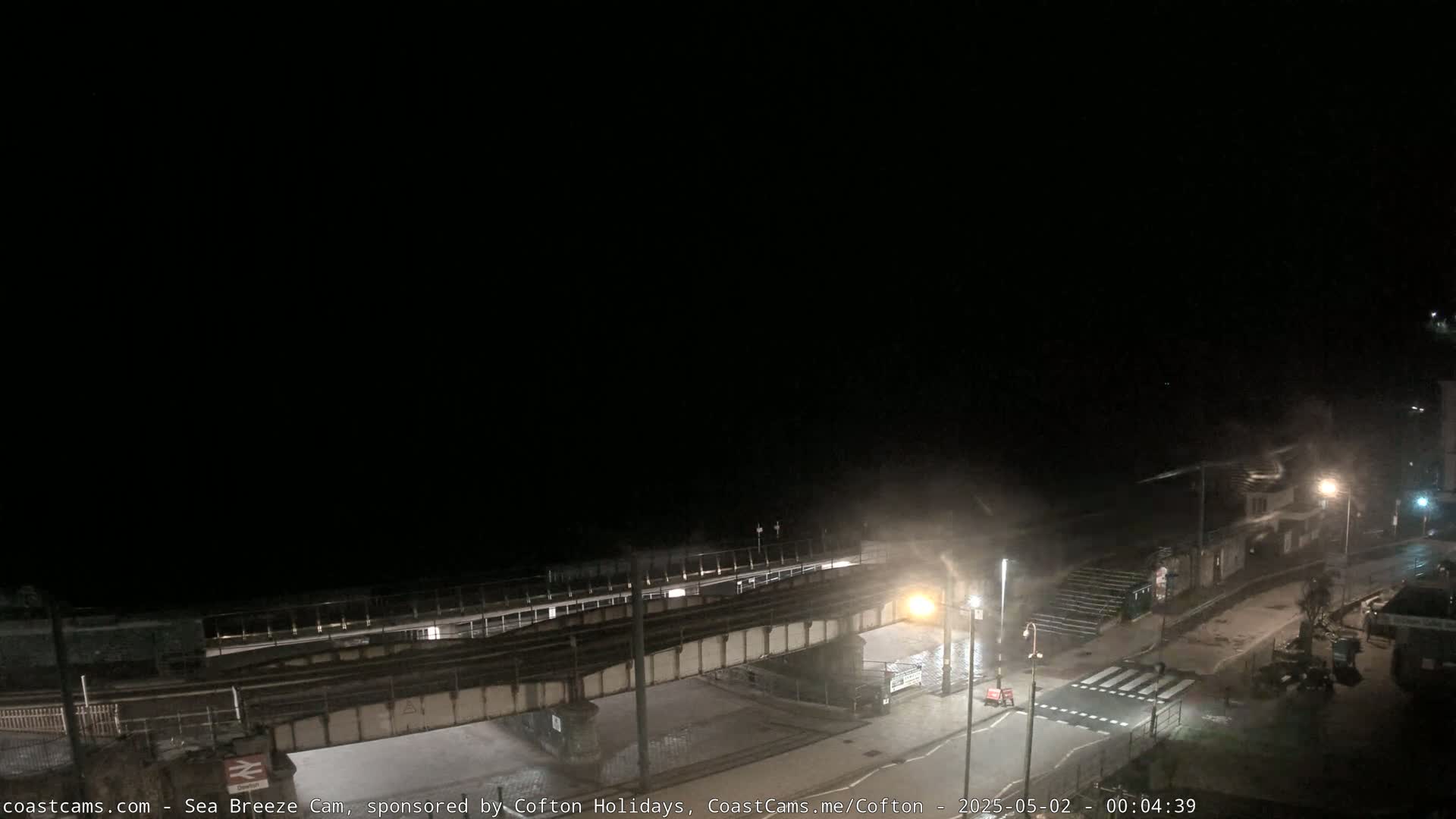 A nighttime, elevated view of a coastal area showing a railway bridge over a road near a beach, with some wind and mist visible.