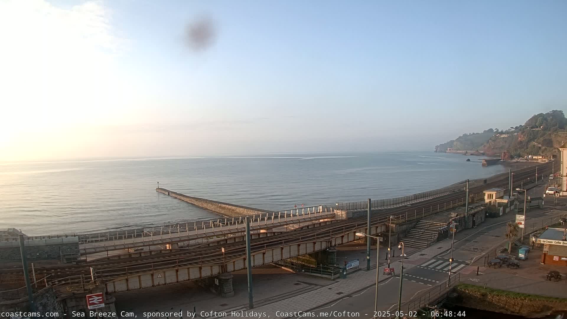 A calm, hazy sunrise illuminates a coastal scene featuring a railway line crossing a road next to a sea wall and a beach, with a town visible on a hillside in the distance.