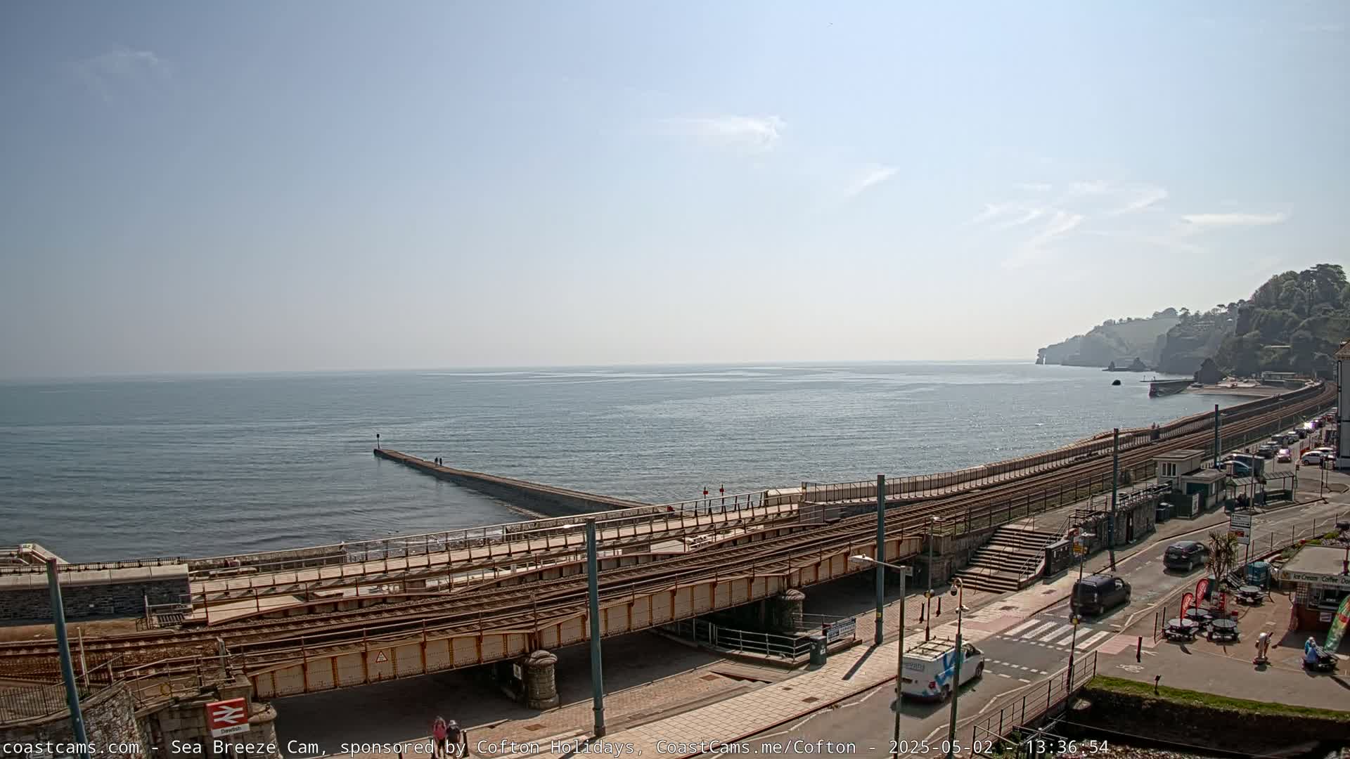 A coastal scene on a sunny day shows a railway track running along a seawall, with a calm ocean and a rocky cliff in the background.