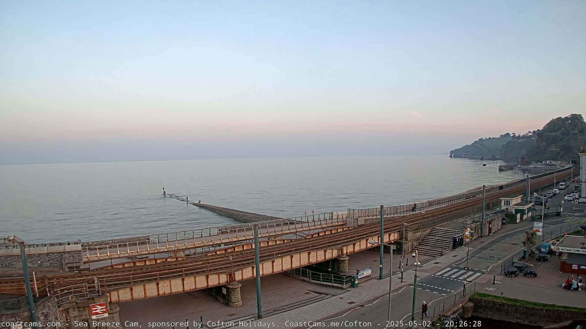 A railway line crosses a coastal road and leads to a sea wall under a calm, twilight sky.