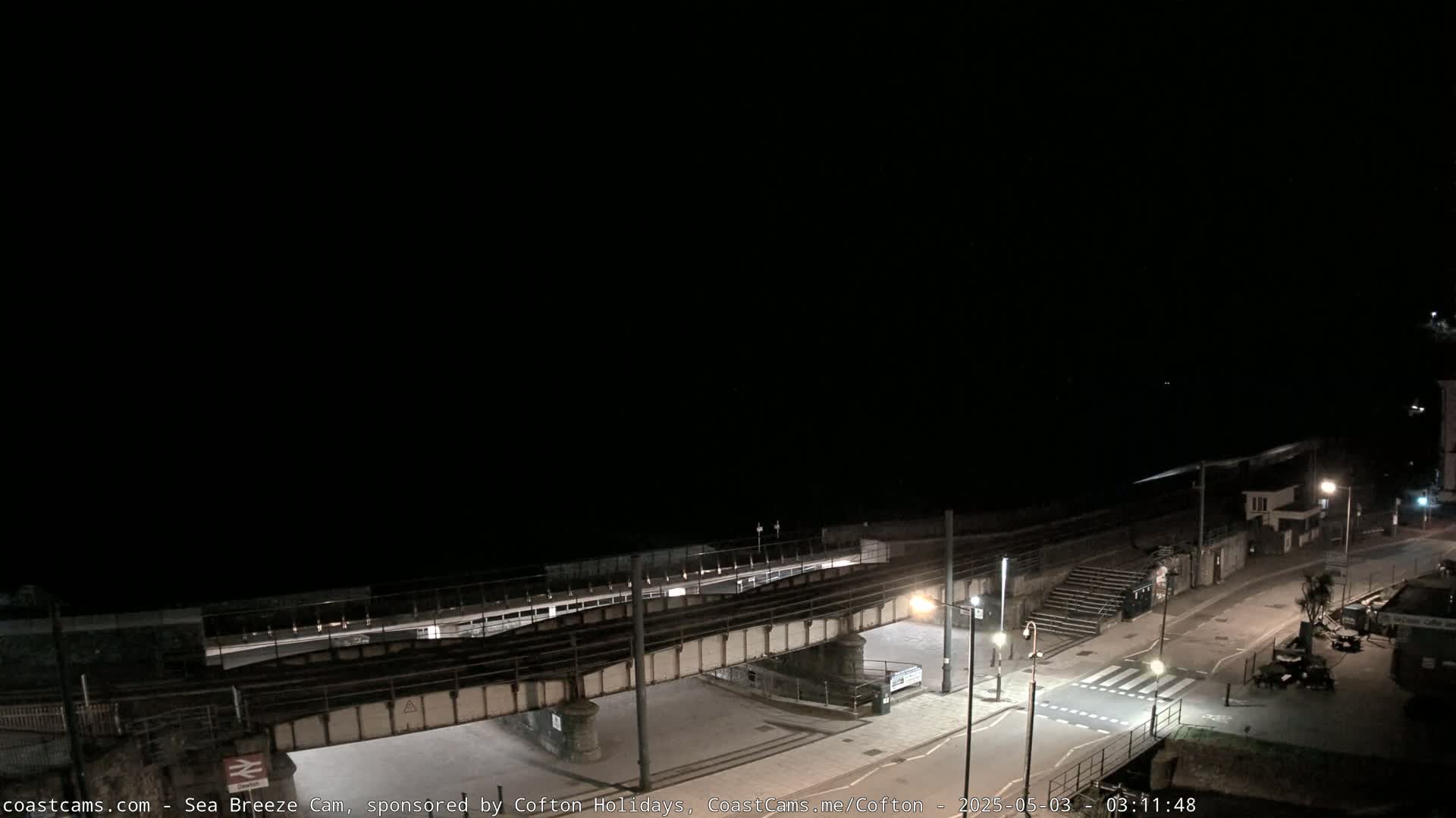A nighttime coastal scene shows a train track overpass above a road and pedestrian crossing, with a dark ocean in the background under a clear, black sky.