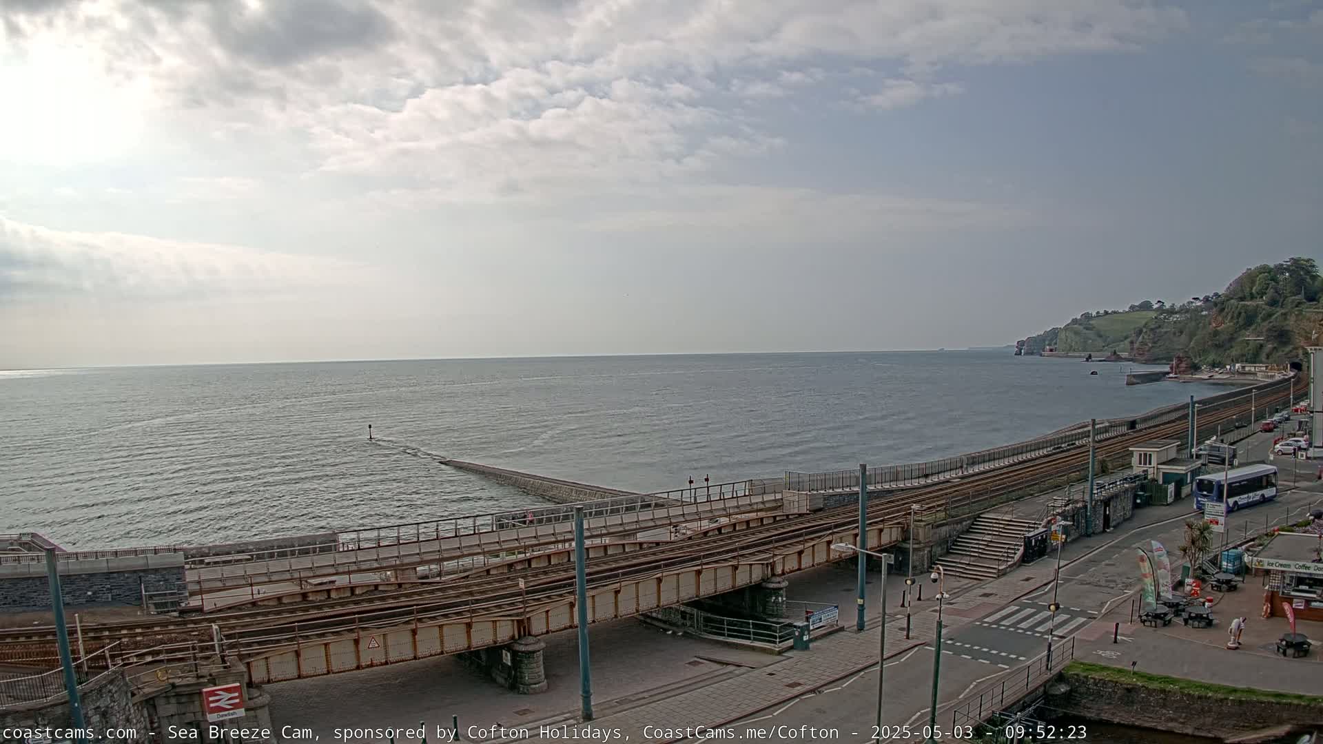 A mostly cloudy day shows a coastal town with train tracks leading to a pier extending into a calm sea, and cliffs visible in the distance.