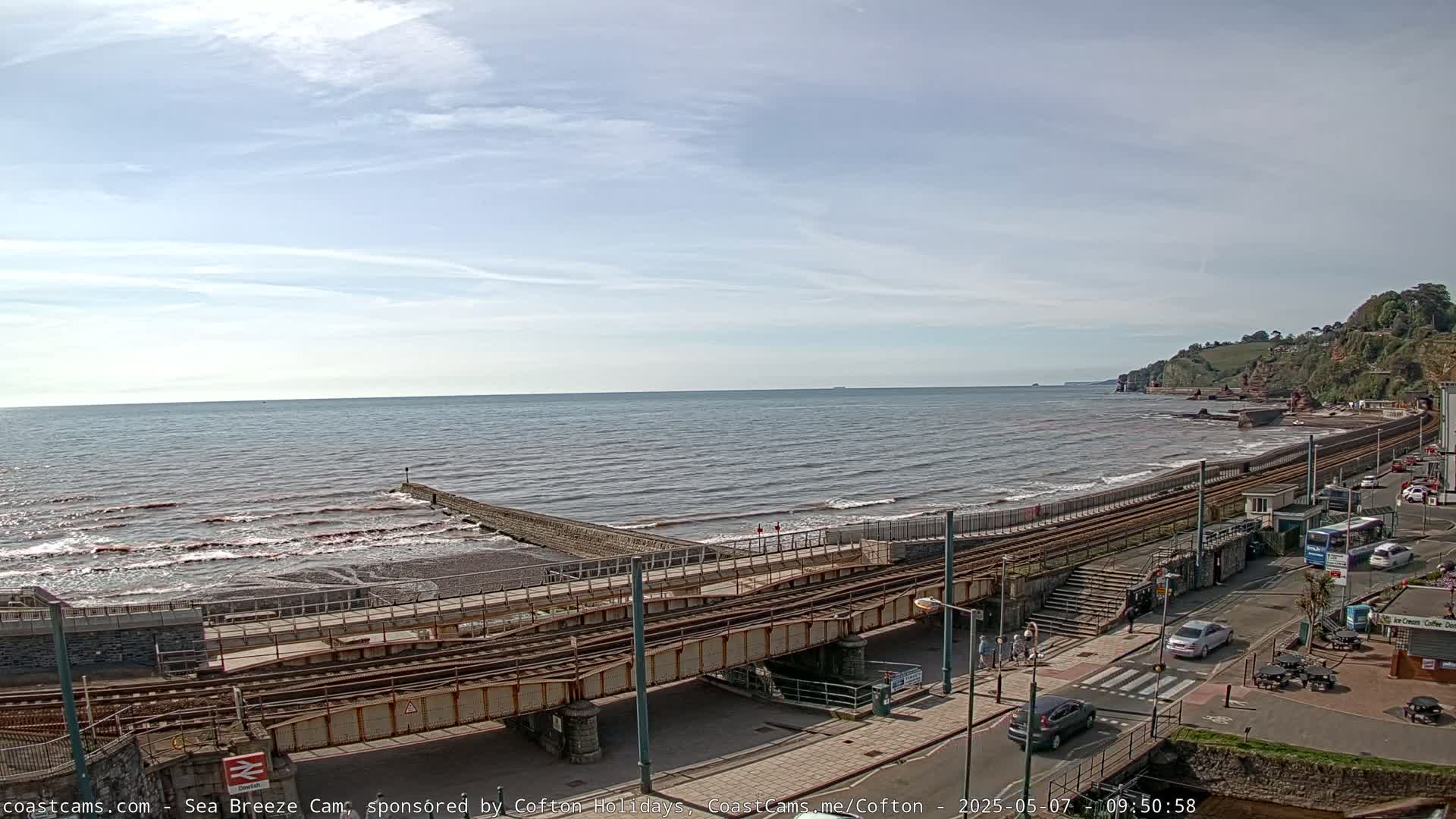 A mostly sunny day shows a coastal scene with a pebble beach, ocean waves, train tracks running alongside the shore, and a road with cars parked beside buildings.