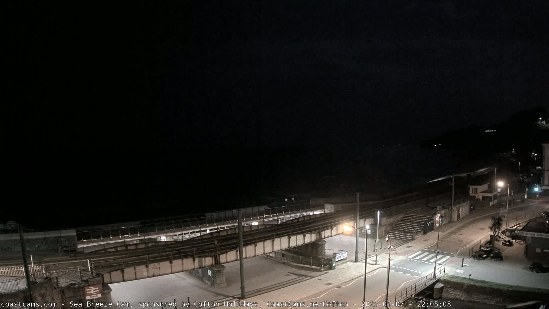 A nighttime view of a coastal town, showing a railway line crossing a road above a beach, under a dark sky.