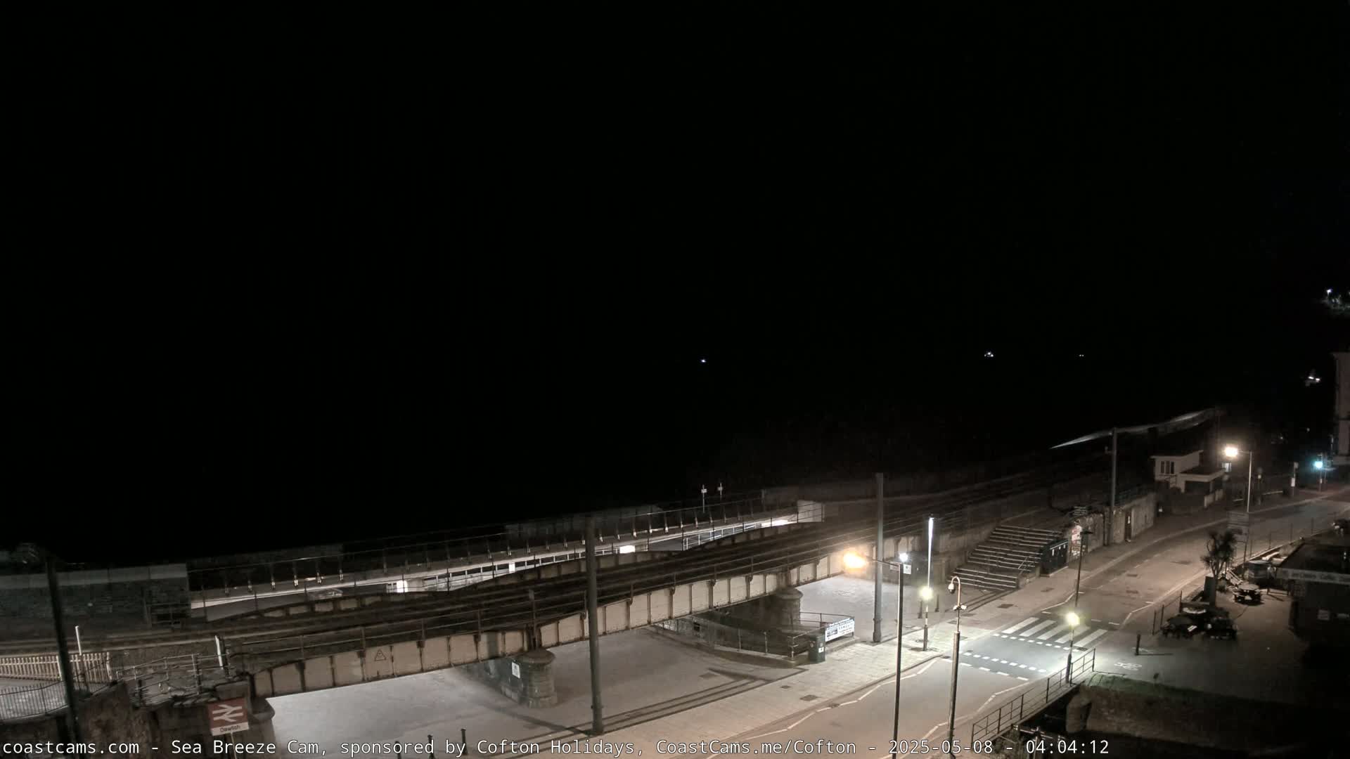 A nighttime view of a coastal town, showing a dark sea, a railway bridge over a road, and sparsely lit streets.