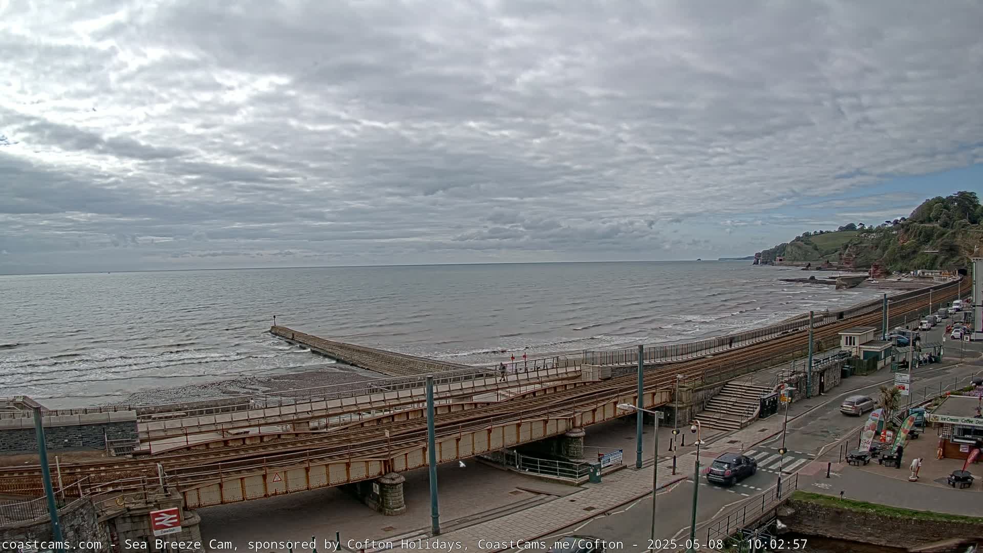 A coastal scene under a cloudy sky shows a railway line crossing a beach with small waves, leading to a town with buildings and vehicles.