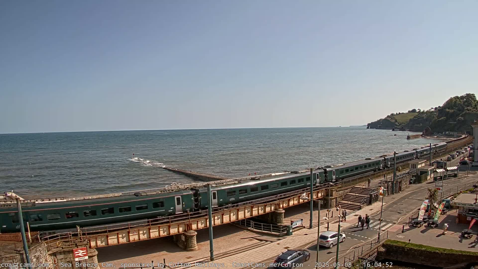 A long, dark-green passenger train travels along a coastal railway line on a sunny day, next to a calm sea.
