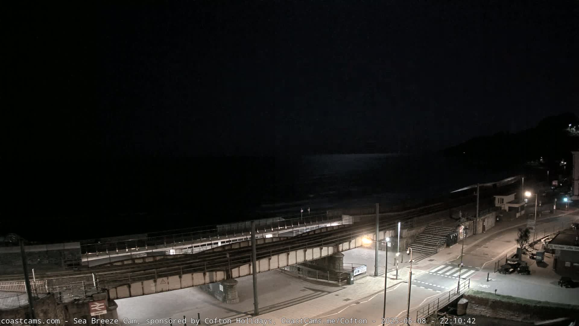 A nighttime view of a coastal town with a railway bridge crossing over a roadway near the ocean, under a dark sky.