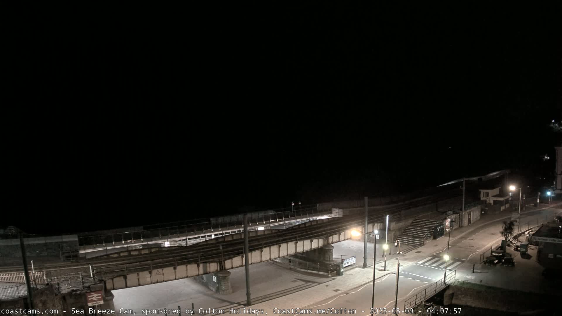 A nighttime, overhead view shows a coastal town with a railway bridge crossing a road near a beach under a dark sky.