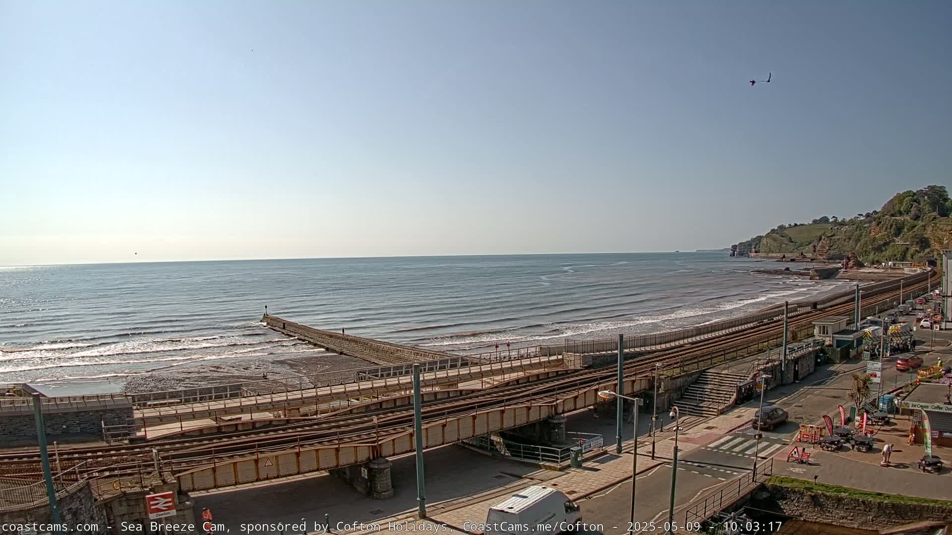 A mostly sunny day shows a coastal scene with gentle waves lapping a beach next to train tracks and a roadway, with cliffs visible in the distance.