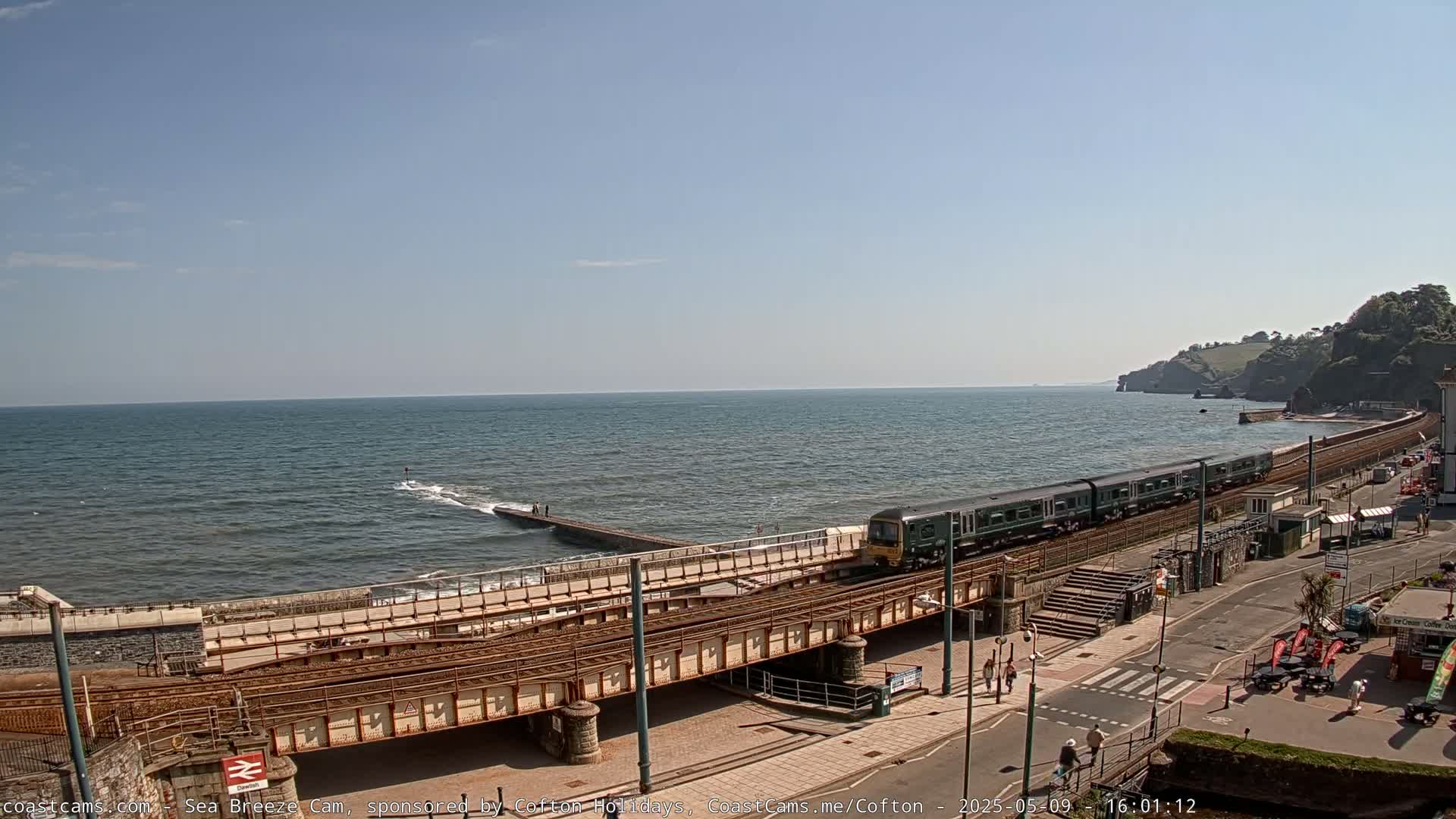 A green passenger train travels across a coastal railway bridge on a sunny day with a calm sea.