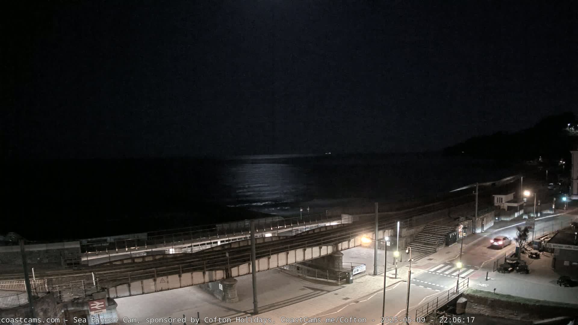 A nighttime coastal scene shows a dark ocean with a faint moon reflection, a railway bridge over a road near a beach, and a few cars on the road.