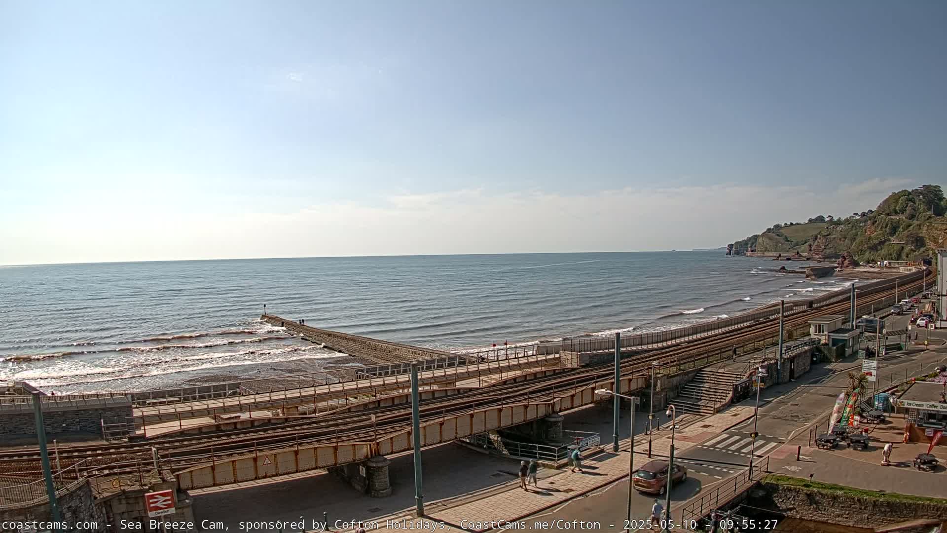 A mostly sunny day shows a rocky beach with gentle waves, a railway line running along the shore, and a town beyond.