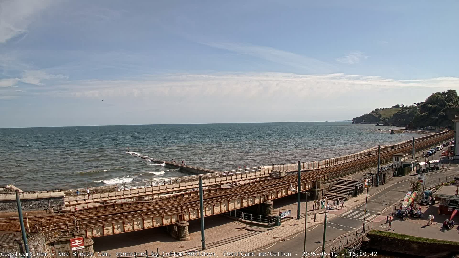 A coastal scene on a sunny day shows a railway line running alongside a seawall, with people walking along the promenade and small waves breaking on the shore.