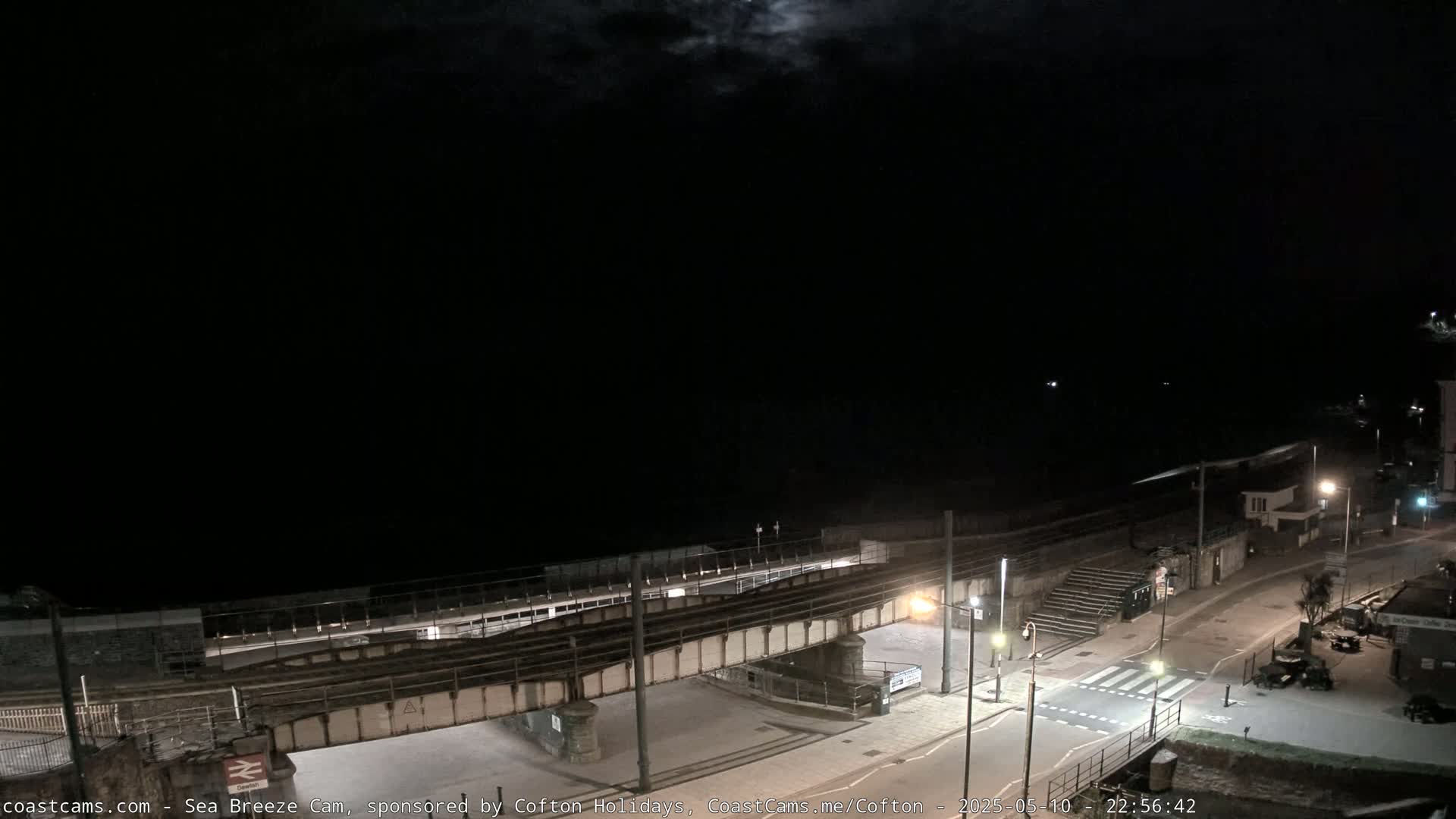 A nighttime coastal scene shows a train track crossing a road near a beach under a partly cloudy moonlit sky.