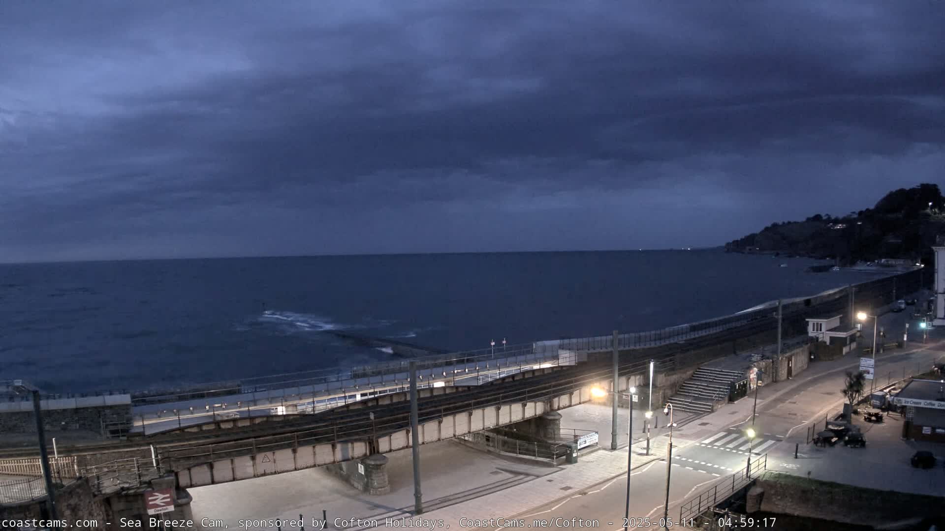 A dark, overcast sky hangs over a calm sea, overlooking a coastal town with train tracks and a road leading to the water.