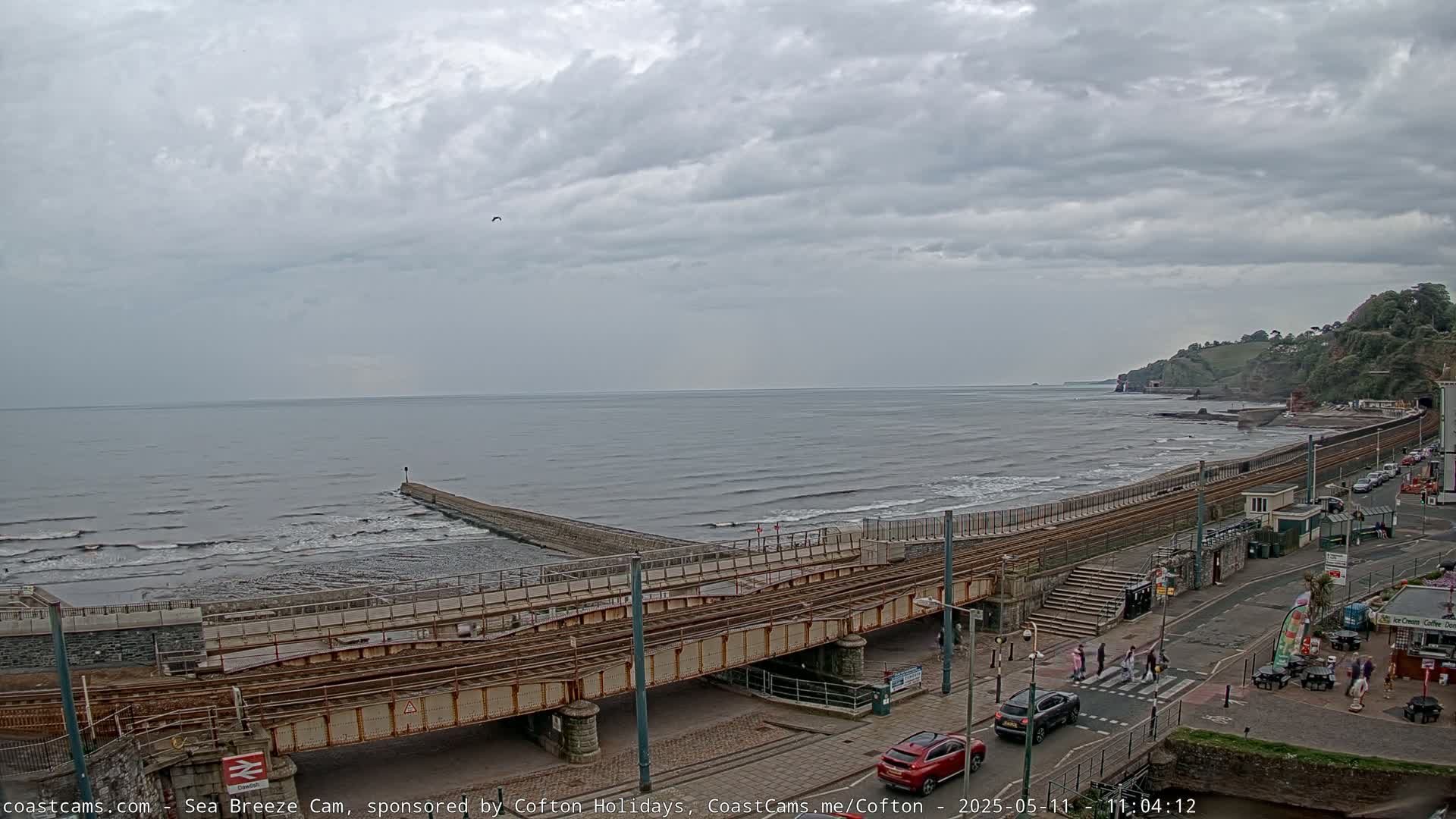A train track crosses a coastal road above a beach with gentle waves under an overcast sky.