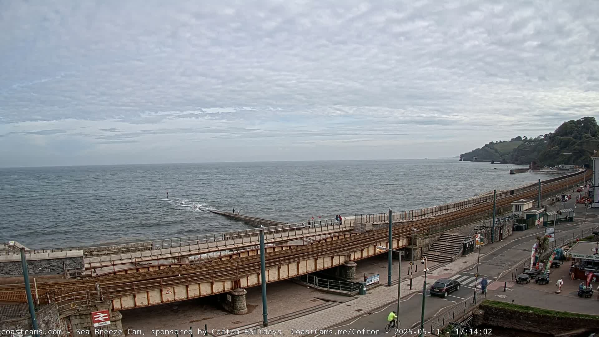 A seaside town scene under a cloudy sky shows a railway line running along a seawall next to a road with some pedestrians and vehicles.