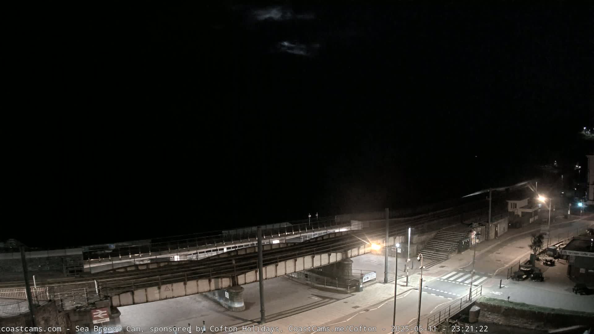 A nighttime view of a coastal railway crossing over a road, under a dark sky with a few visible clouds.