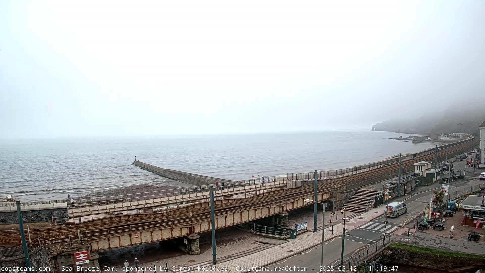 A foggy coastal scene shows a railway bridge crossing a beach leading to a calm sea and a distant, fog-covered hillside.