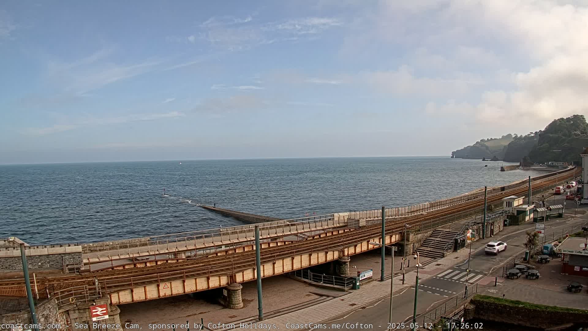 A coastal scene on a partly cloudy day shows a railway track extending along a sea wall, with a calm ocean and a distant shoreline.