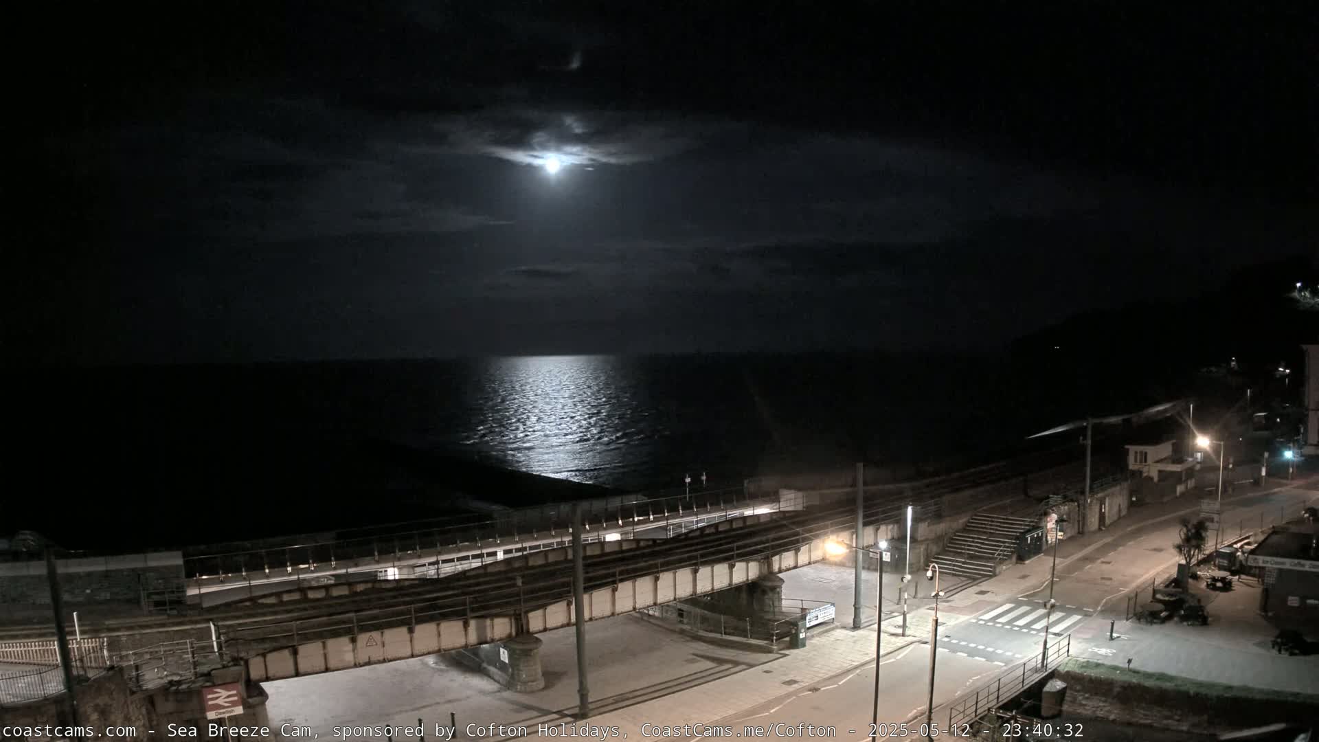 A moonlit night scene overlooking a calm sea, with a railway crossing and street visible below.