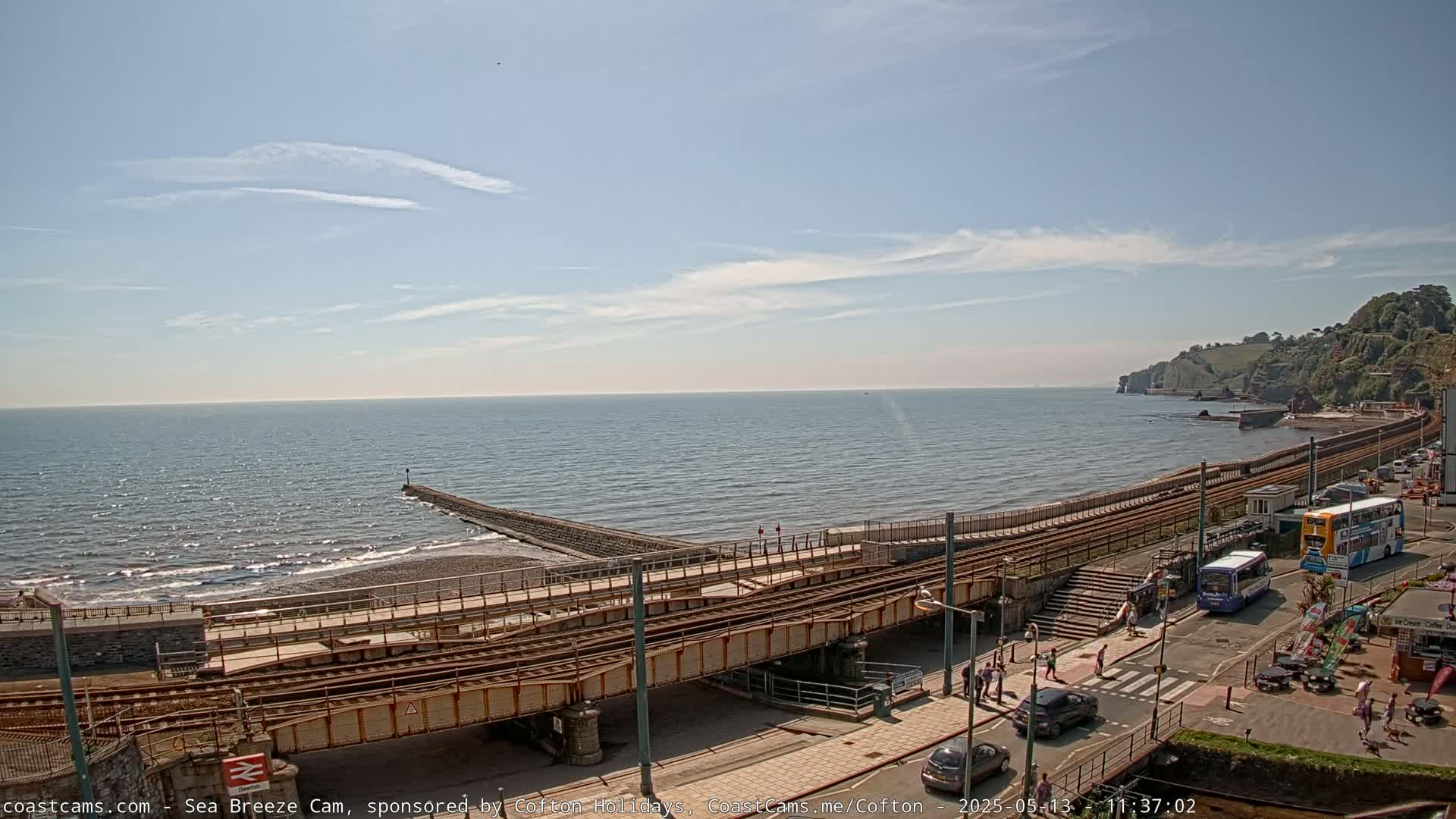 A mostly sunny day shows a coastal scene with a train track running along a pebble beach next to a calm ocean and a small town on a cliff in the distance.