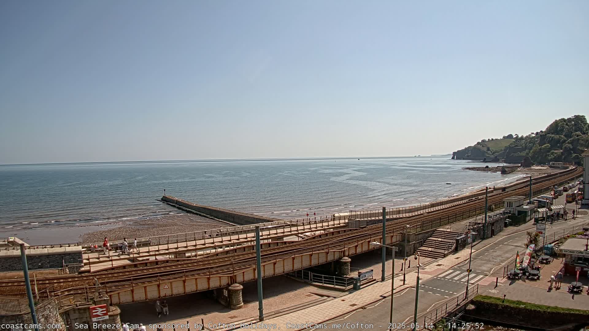 A seaside scene on a sunny day shows a beach with a railway line running along it, leading to a coastal town with green hills in the background.