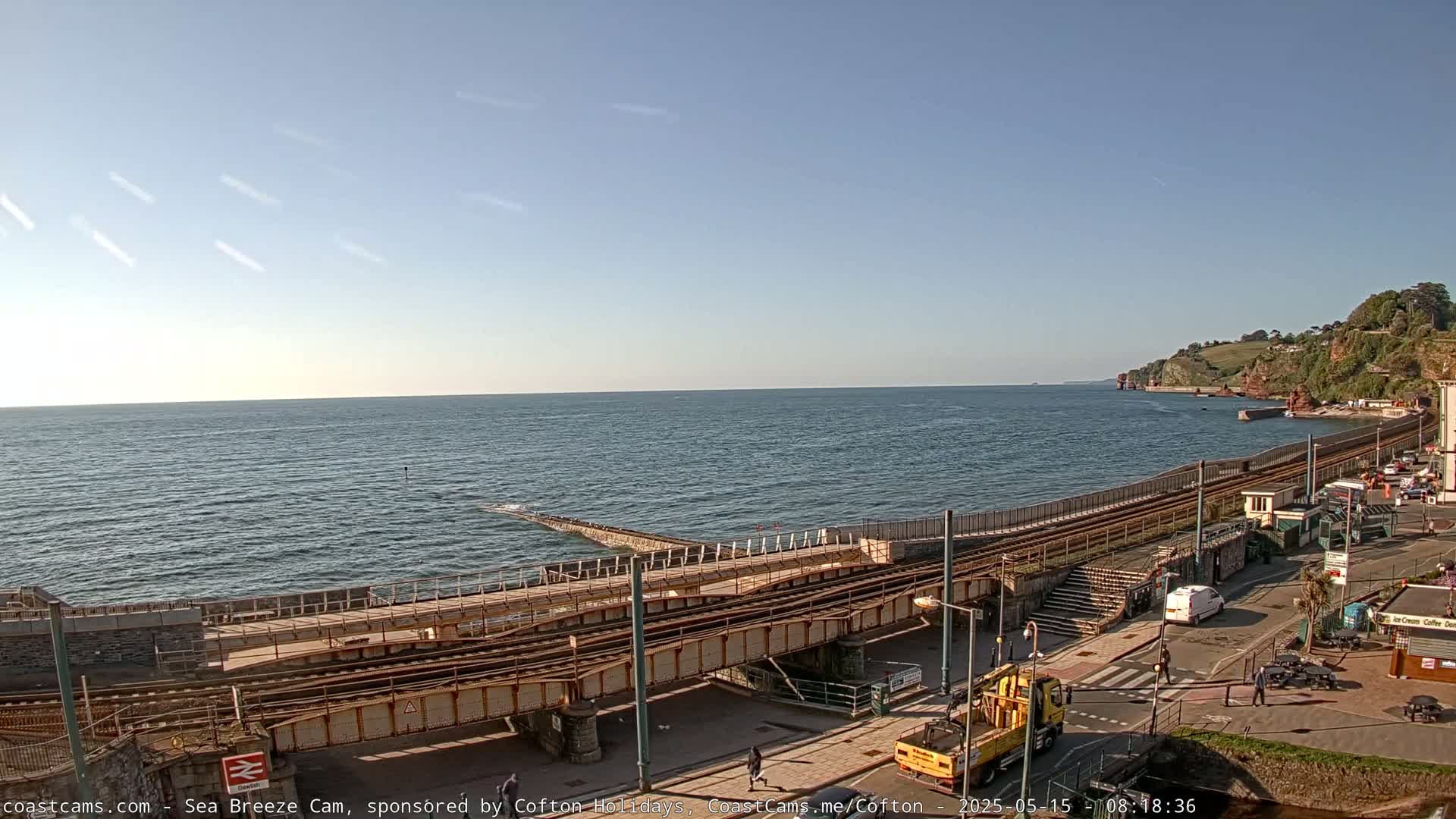 A coastal scene under a clear sky shows a train track leading to a seawall, with a calm sea extending to the horizon and a small town visible along the shore.
