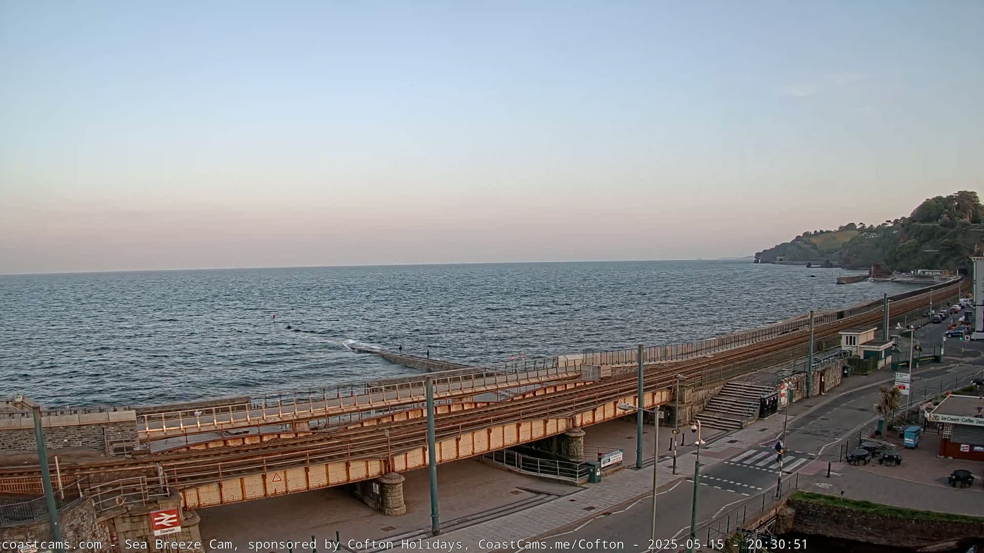 A coastal scene at dusk shows a railway line running along a seawall next to a road, with a choppy sea beyond.