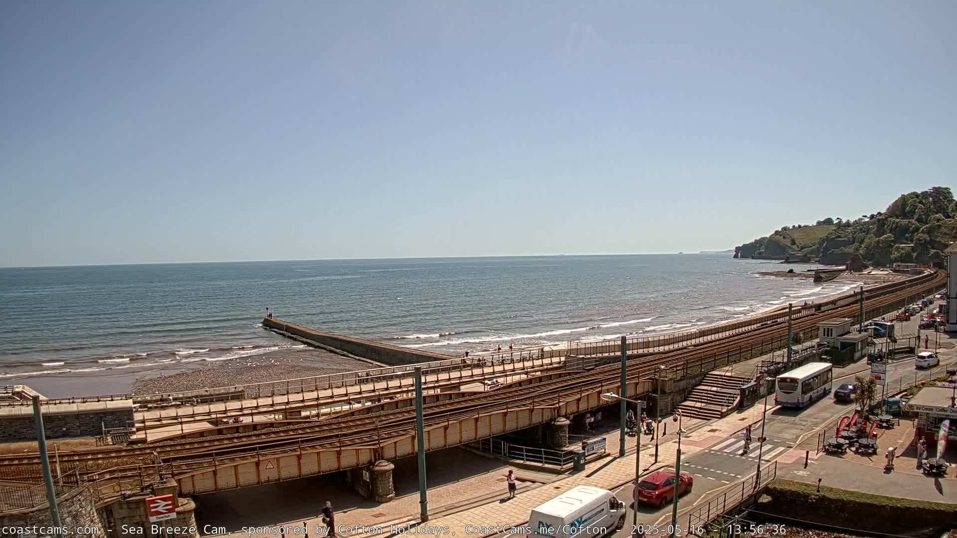A seaside scene on a sunny day shows a beach with a railway line running along it, next to a road with vehicles, and a small town on a hillside in the distance.