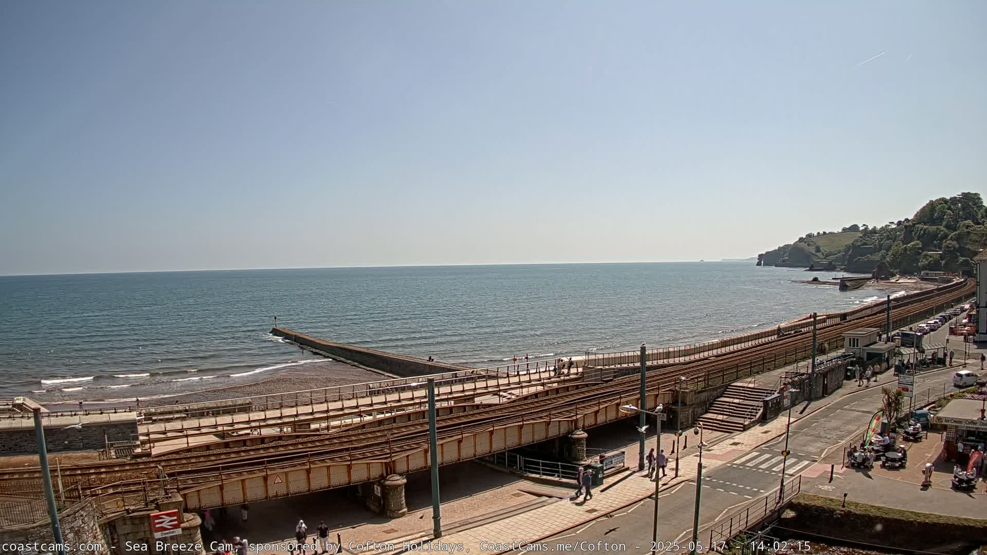 A railway line runs alongside a beach and calm ocean under a clear blue sky.