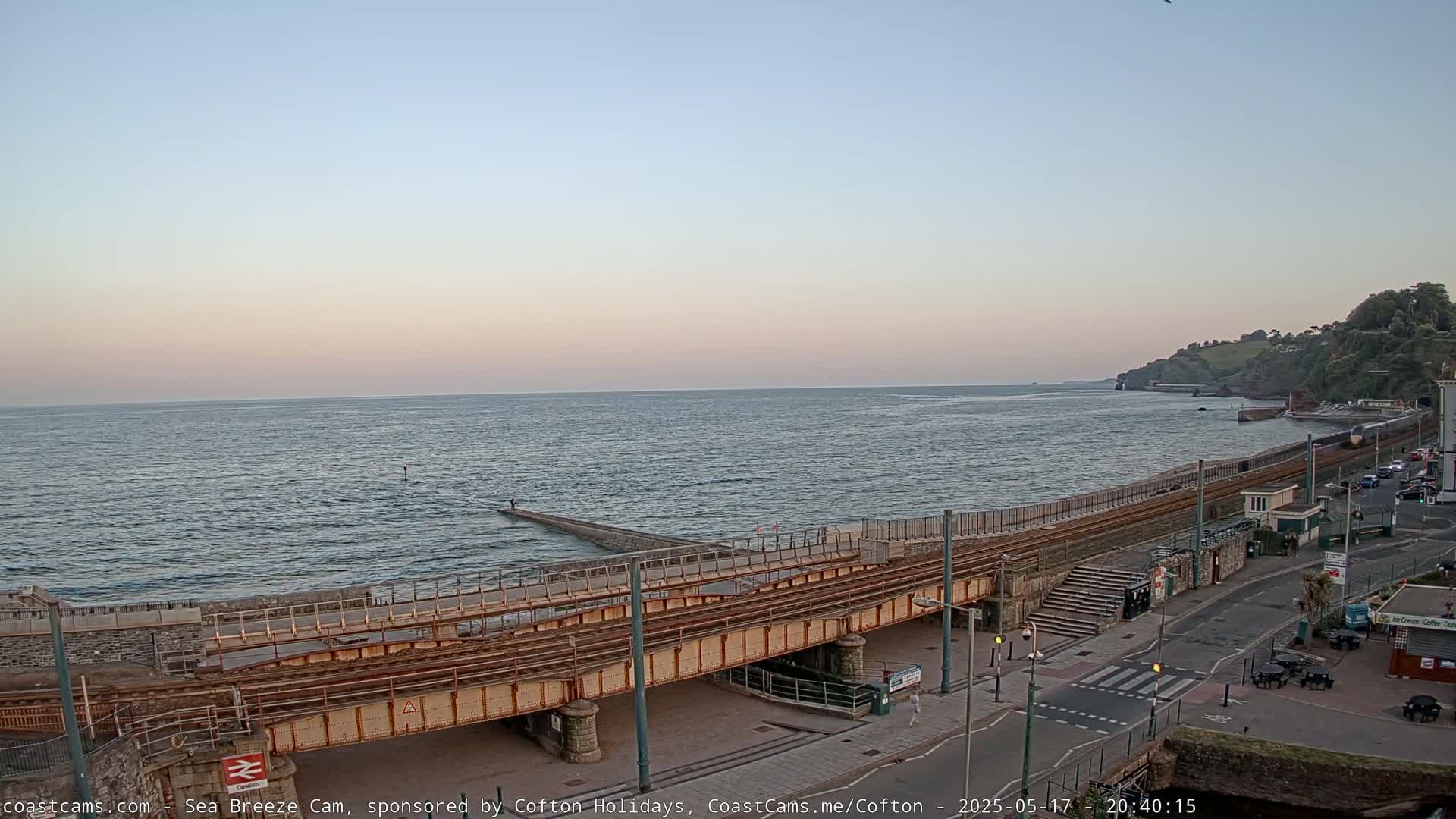A coastal scene at dusk shows a train track crossing a low stone wall and leading to a roadway, with a calm sea extending to a distant shoreline under a pale sky.