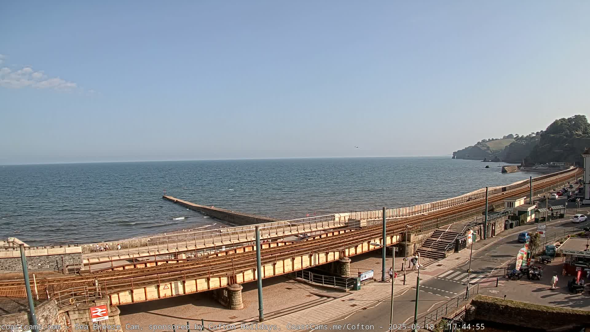 A railway line runs along a sea wall next to a beach, with a calm sea under a clear blue sky.